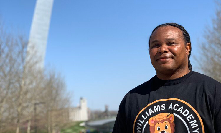 A person with braided hair stands outdoors wearing a "Williams Academy" shirt with a bear logo. In the background, the St. Louis Gateway Arch rises against a clear blue sky and leafless trees.