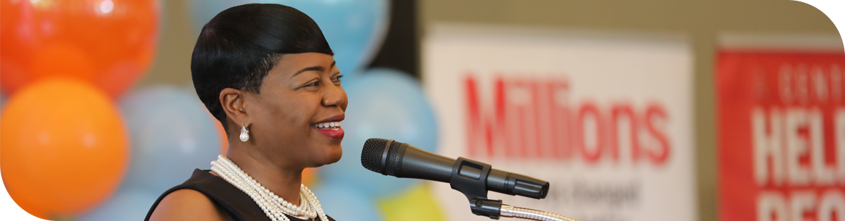 Michelle Tucker, with short black hair and pearl jewelry, smiles while speaking into a microphone. They are standing in front of colorful balloons and banners with text partially visible.